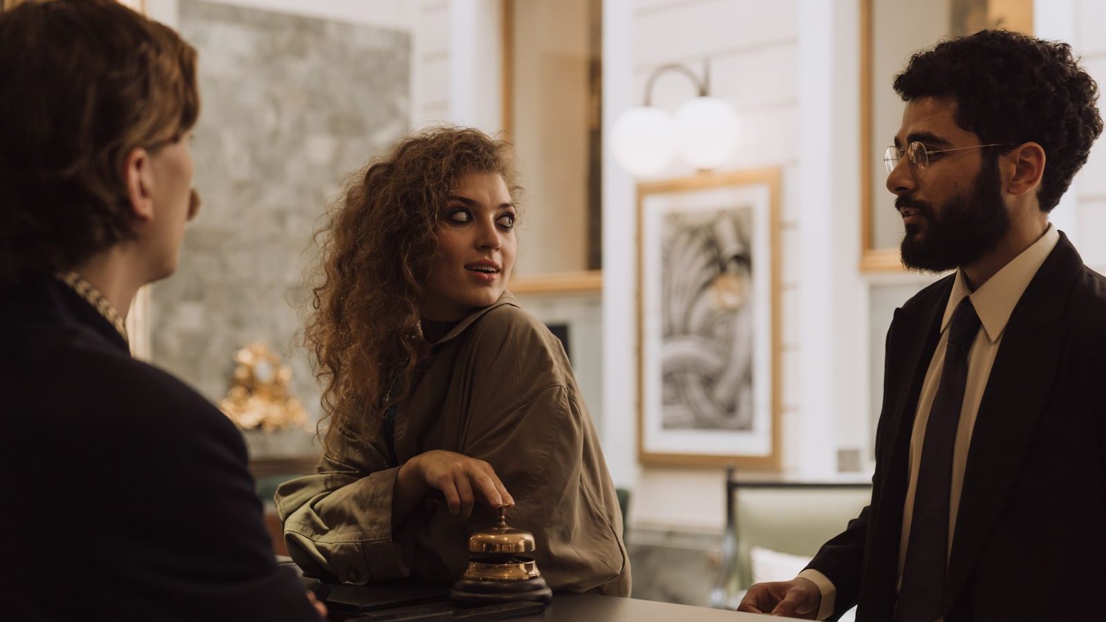 Una mujer conversa en el mostrador de recepción de un hotel con un recepcionista con barba y gafas, mientras otra persona escucha de espaldas.