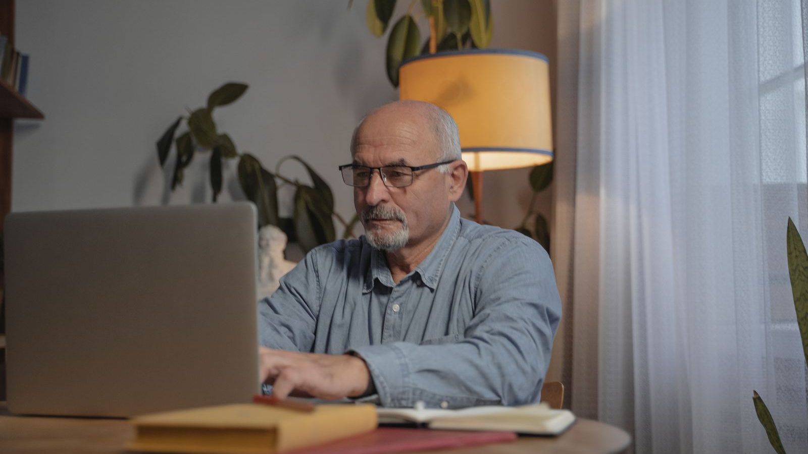 Hombre mayor con gafas escribiendo en su portátil en la mesa de casa, con un cuaderno y una carpeta al lado y una lámpara de pie detrás.